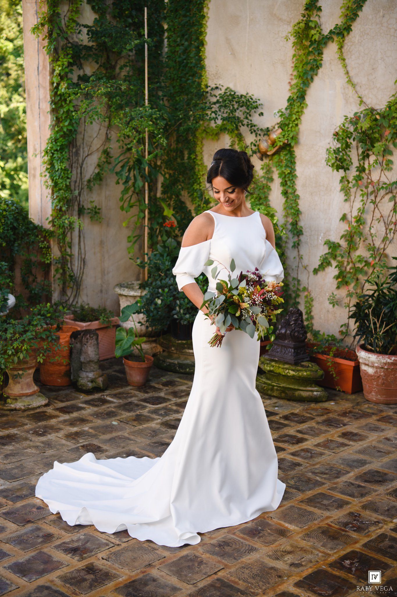 Bride in white off-shoulder mermaid gown holds bouquet, standing on cobblestone patio with ivy-covered walls.