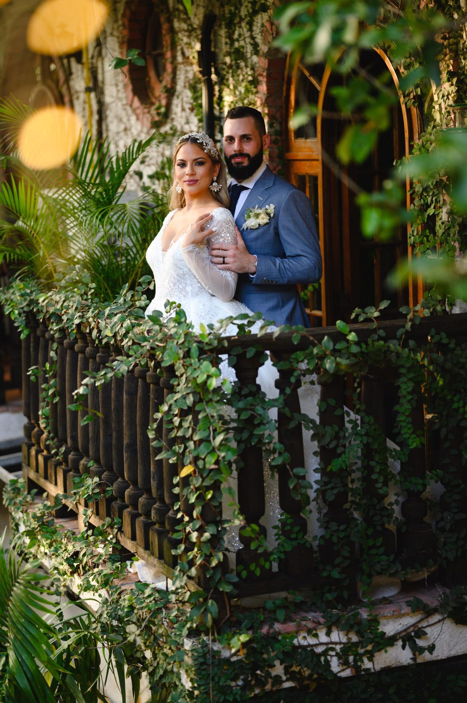 Bride and groom pose on ivy-covered balcony surrounded by lush tropical greenery in elegant wedding attire.