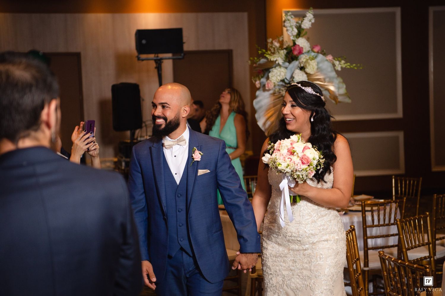 Smiling bride and groom walk hand-in-hand at their wedding reception, bride holding a pink bouquet.