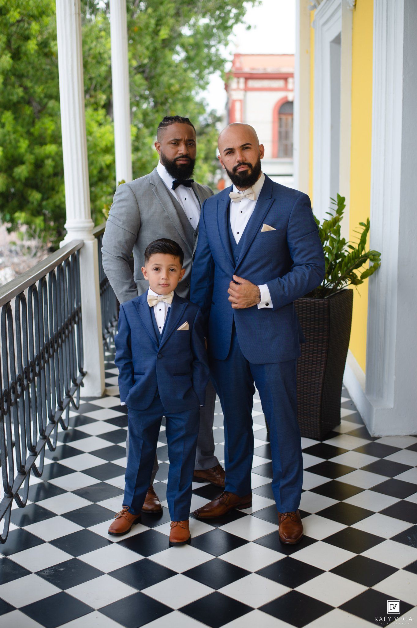 Two men and a boy in matching blue suits pose on a checkered floor balcony at a wedding.