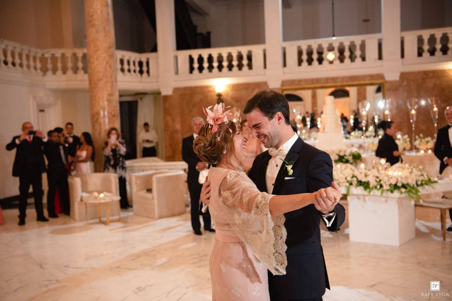 Bride and groom share their first dance in an elegant ballroom with marble floors and guests watching in the background.