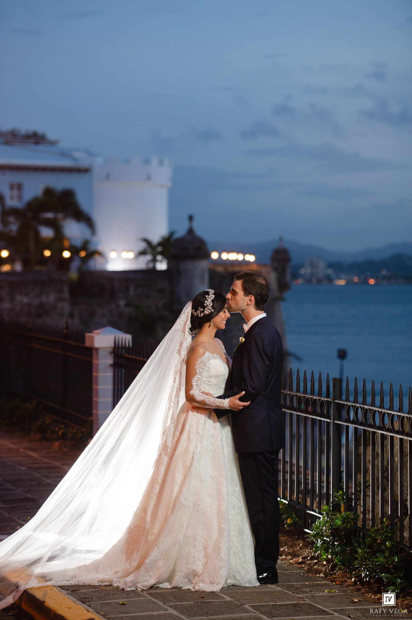 Bride and groom share a romantic kiss at dusk with ocean views and historic architecture in the background.