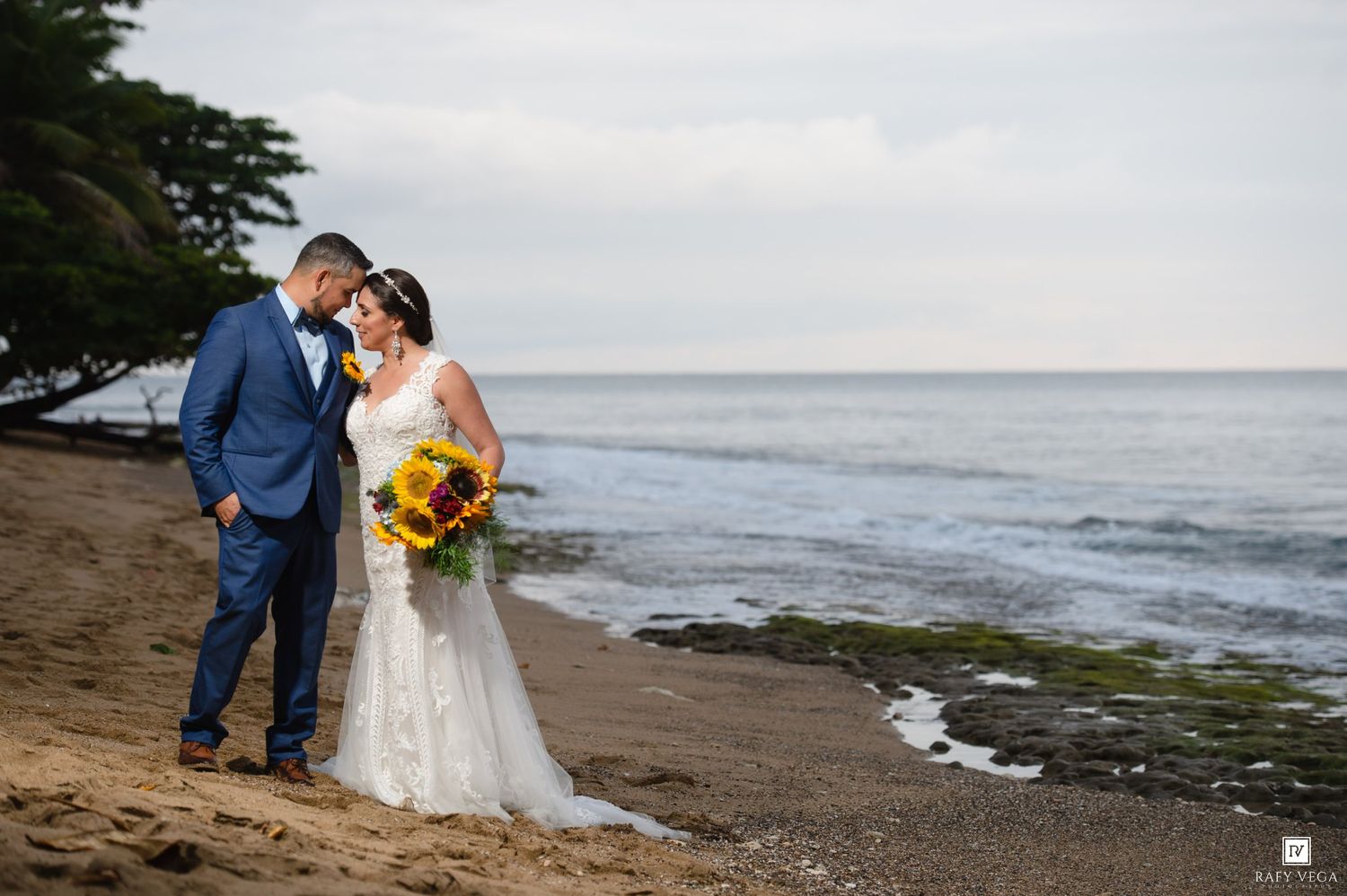 Bride and groom share romantic moment on sandy beach with ocean waves and lush trees behind them.