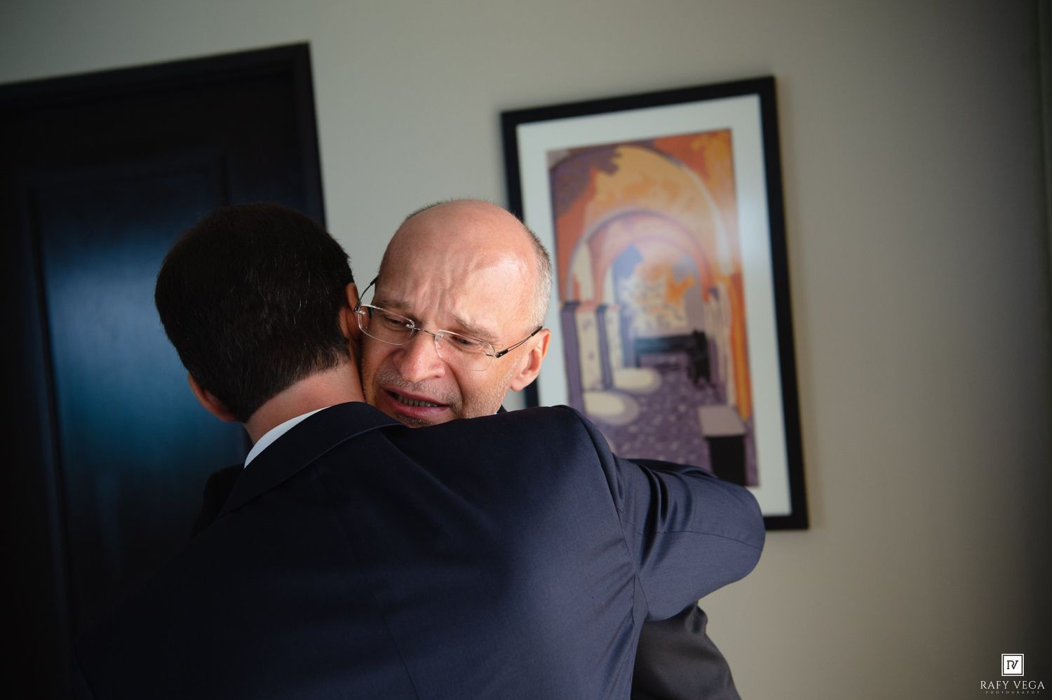 Two men in suits sharing a warm embrace at a wedding celebration in an elegantly decorated room.