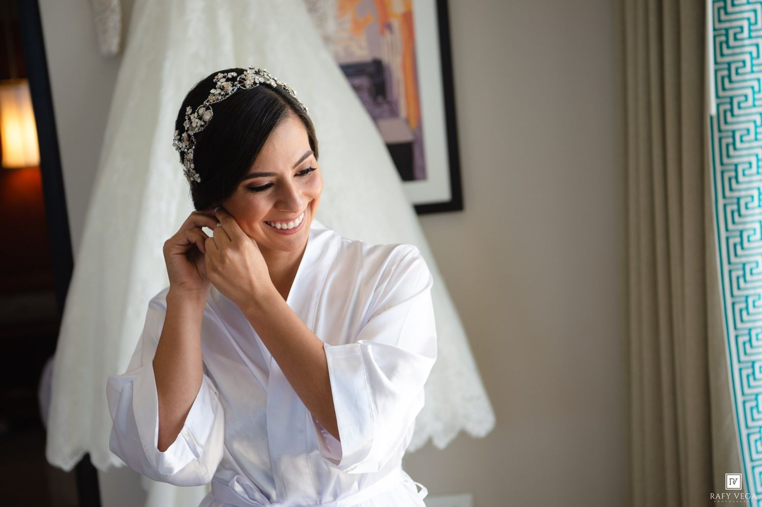 Bride in white robe adjusting earrings with wedding veil and tiara, smiling near window on wedding day.