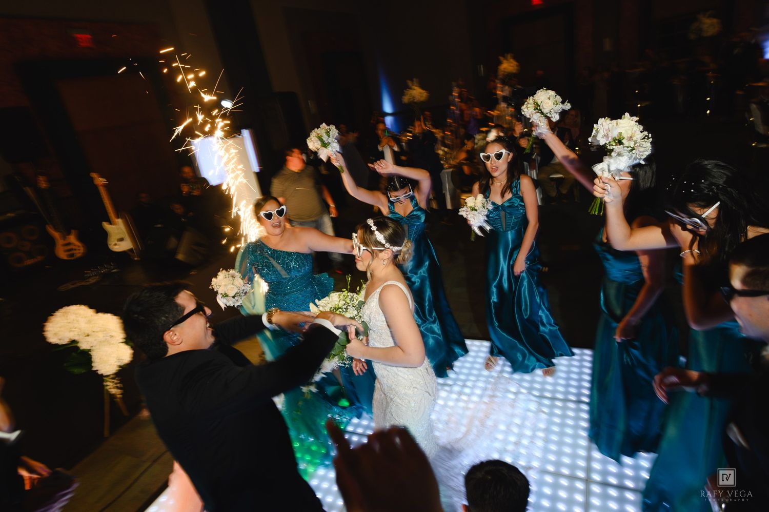 Bridesmaids in teal dresses celebrate with sparklers and bouquets on illuminated dance floor at wedding reception.