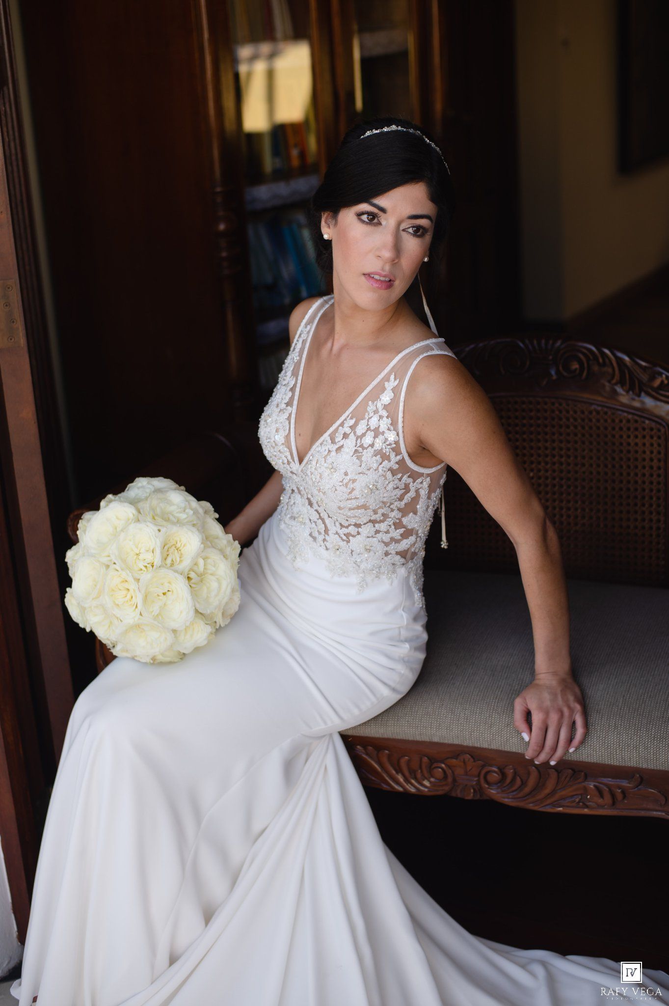 Elegant bride in a lace V-neck wedding gown holding white peony bouquet, seated on ornate wooden bench.