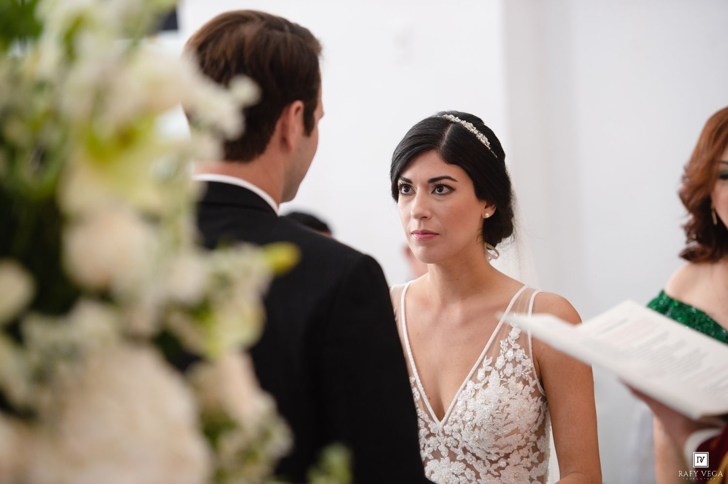 Bride in beaded gown with floral headpiece gazes at groom during elegant wedding ceremony with white floral arrangements.