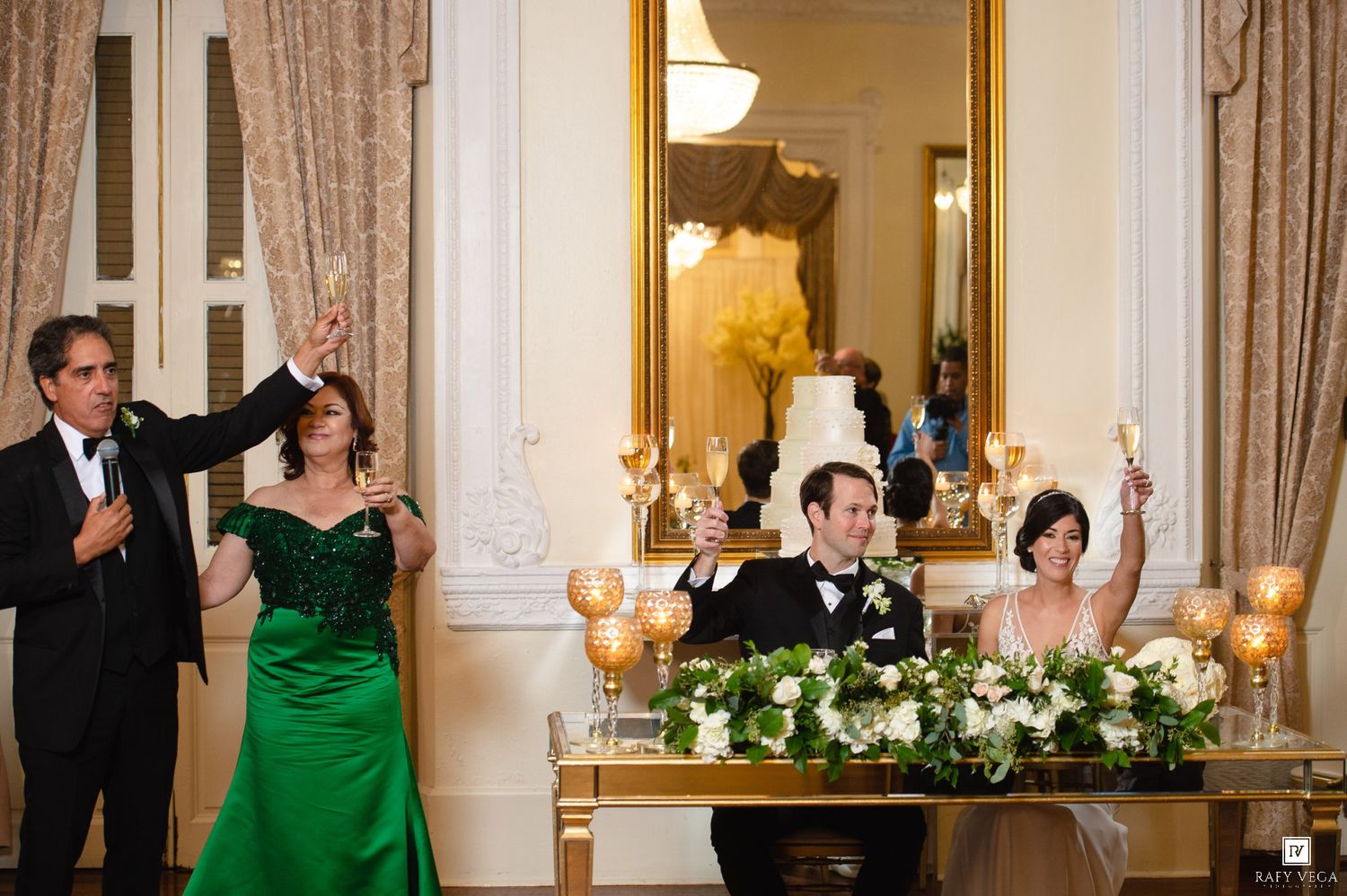 Wedding party raises champagne glasses in a toast at a formal reception with floral arrangements and candlelight.