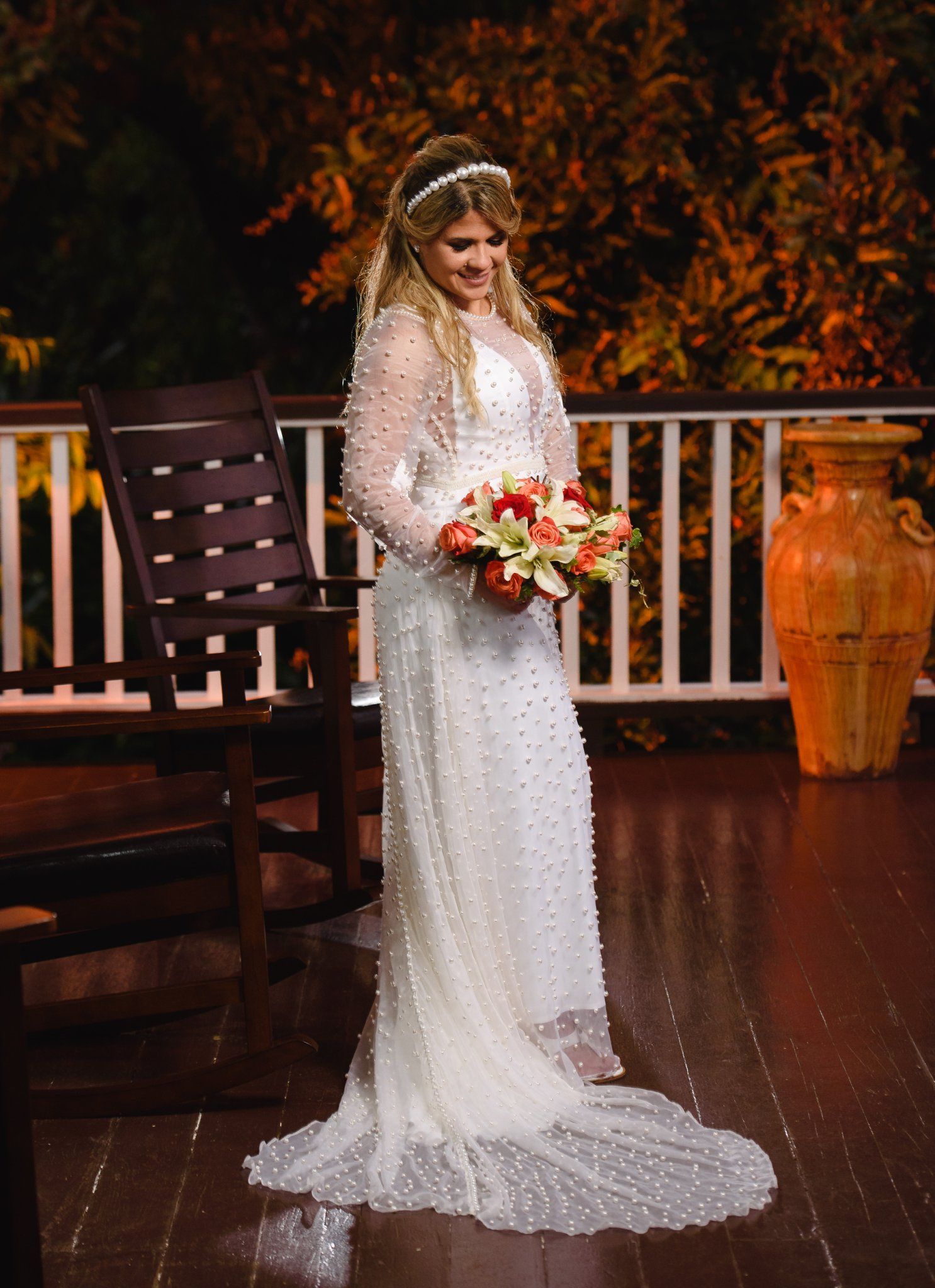Bride in a long-sleeve beaded white wedding gown holding a red bouquet, smiling on a wooden deck at night.