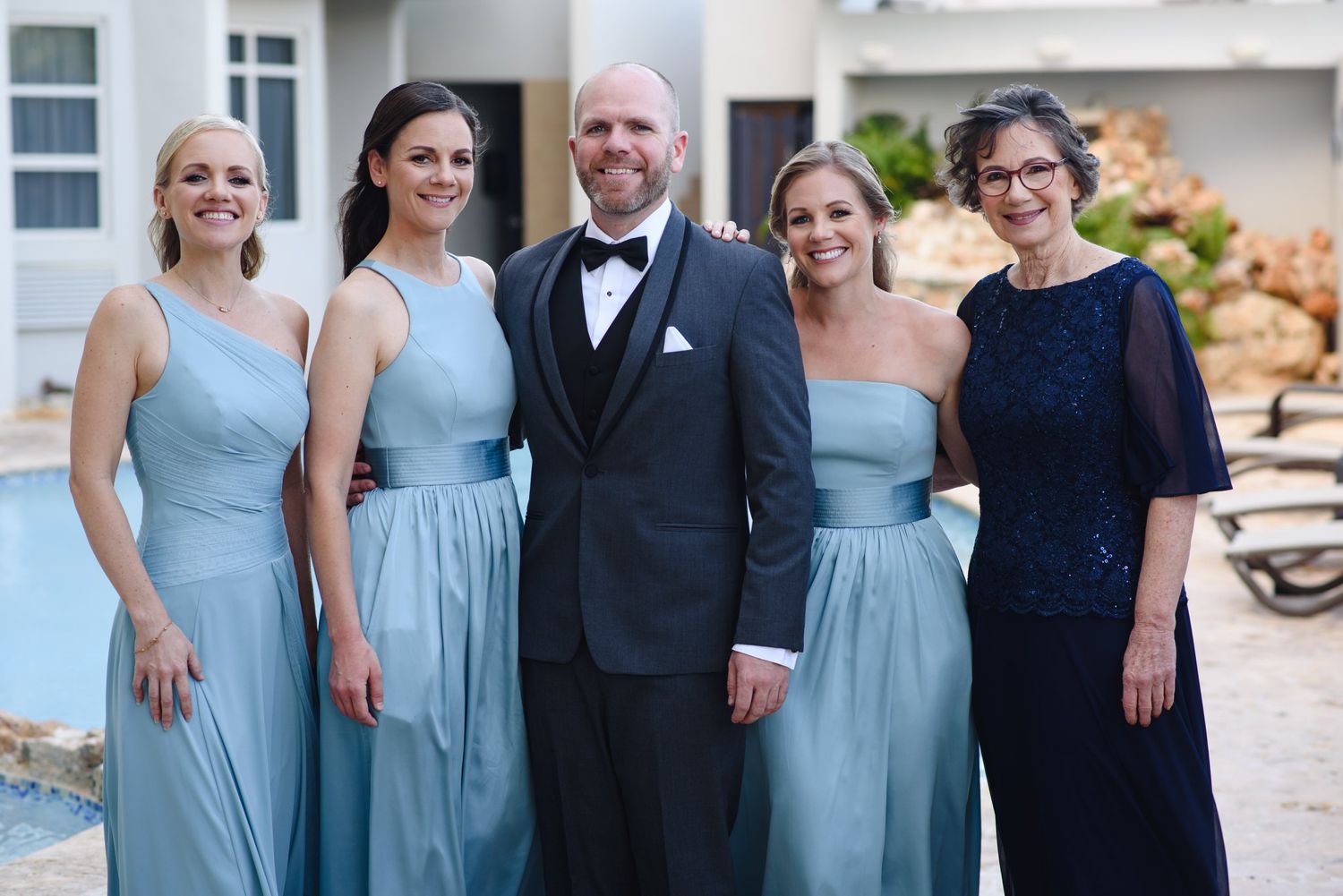 Groom in tuxedo poses with four women in dusty blue bridesmaid dresses and navy gown outdoors.