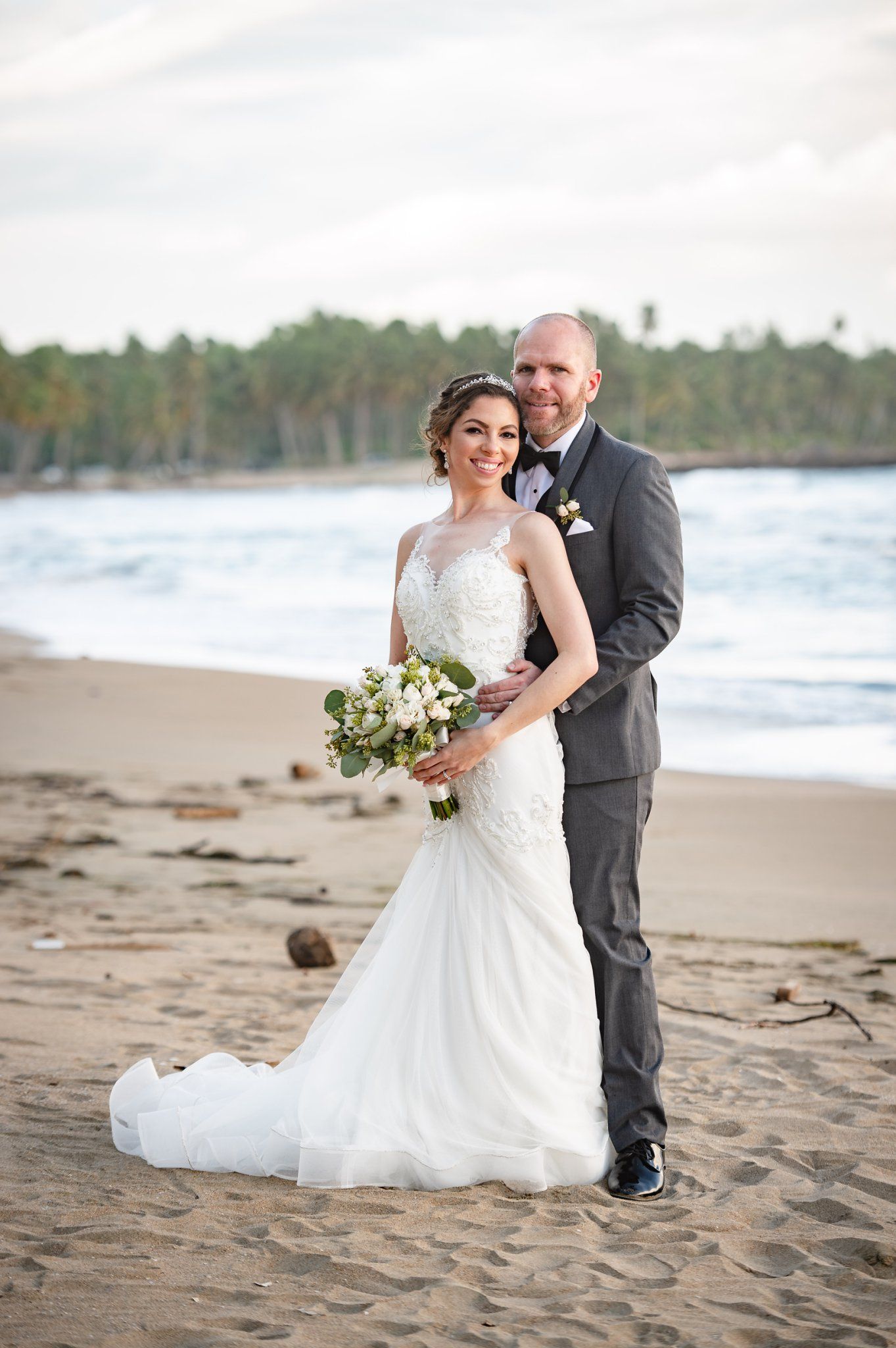 Smiling bride and groom posing together on a tropical beach, bride in lace gown holding a white floral bouquet.