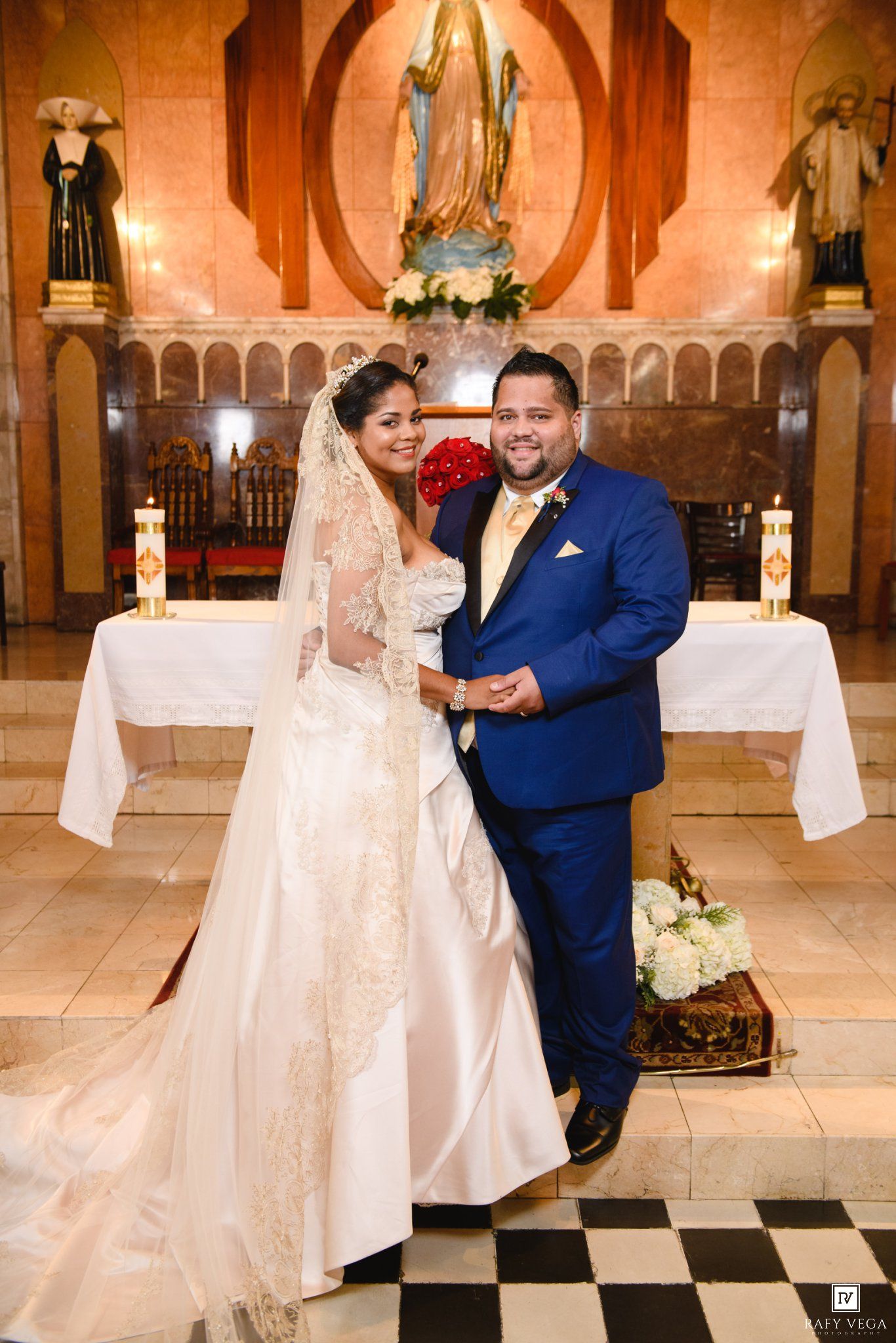 Bride in white gown with veil and groom in blue suit pose together at church altar after wedding ceremony.