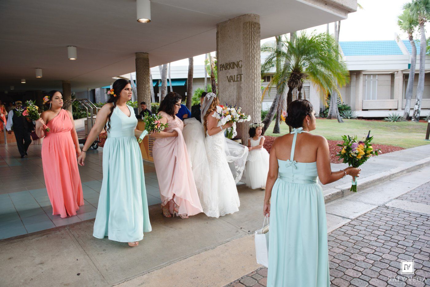 Bridesmaids in mint and coral gowns walk with a bride in white outside a tropical venue with palm trees.
