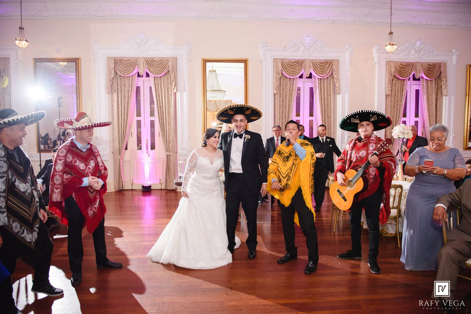 Bride and groom enter wedding reception accompanied by mariachi band in traditional charro suits on dance floor.