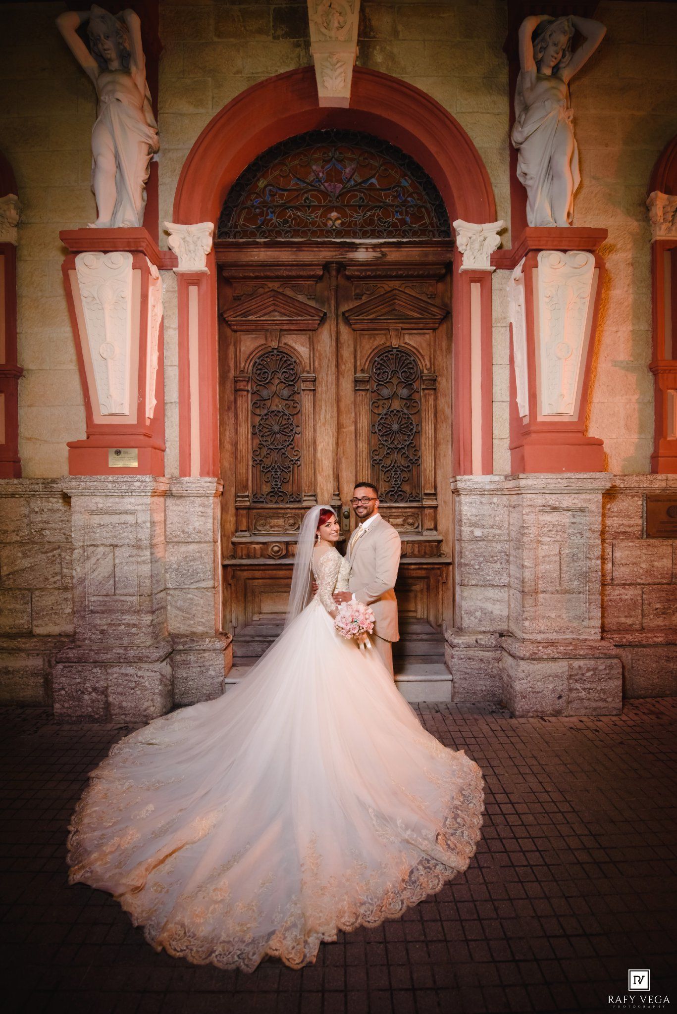 Bride and groom posing in front of ornate wooden doors with stone statues, bride wearing elegant lace gown with long train.