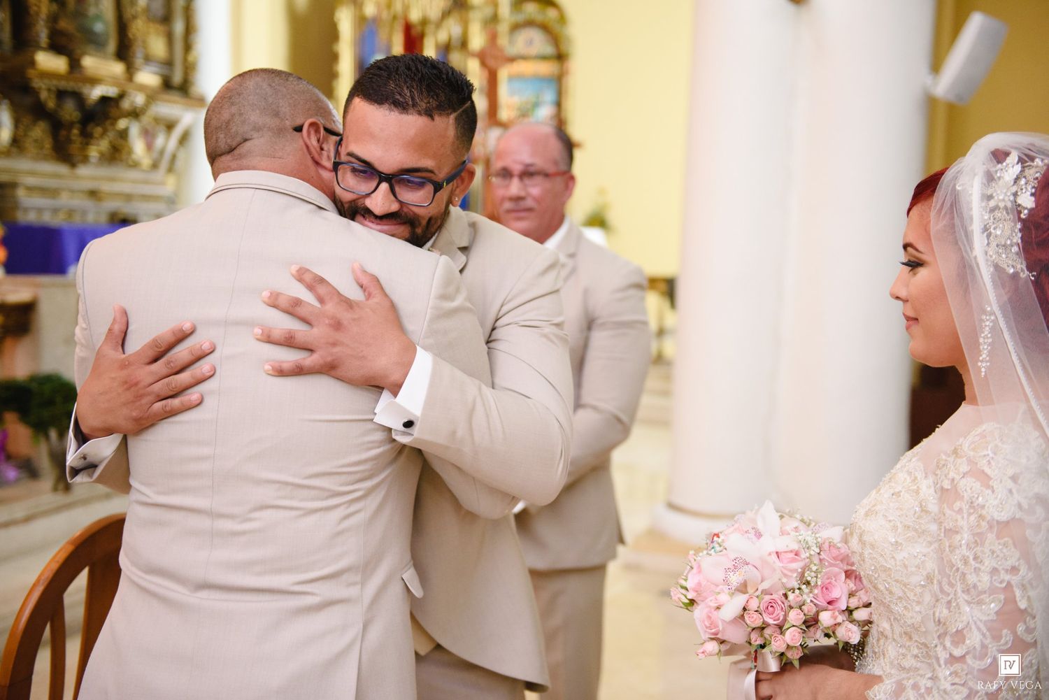 Groom in tan suit embraces guest at church altar while bride in lace gown holds pink bouquet nearby.
