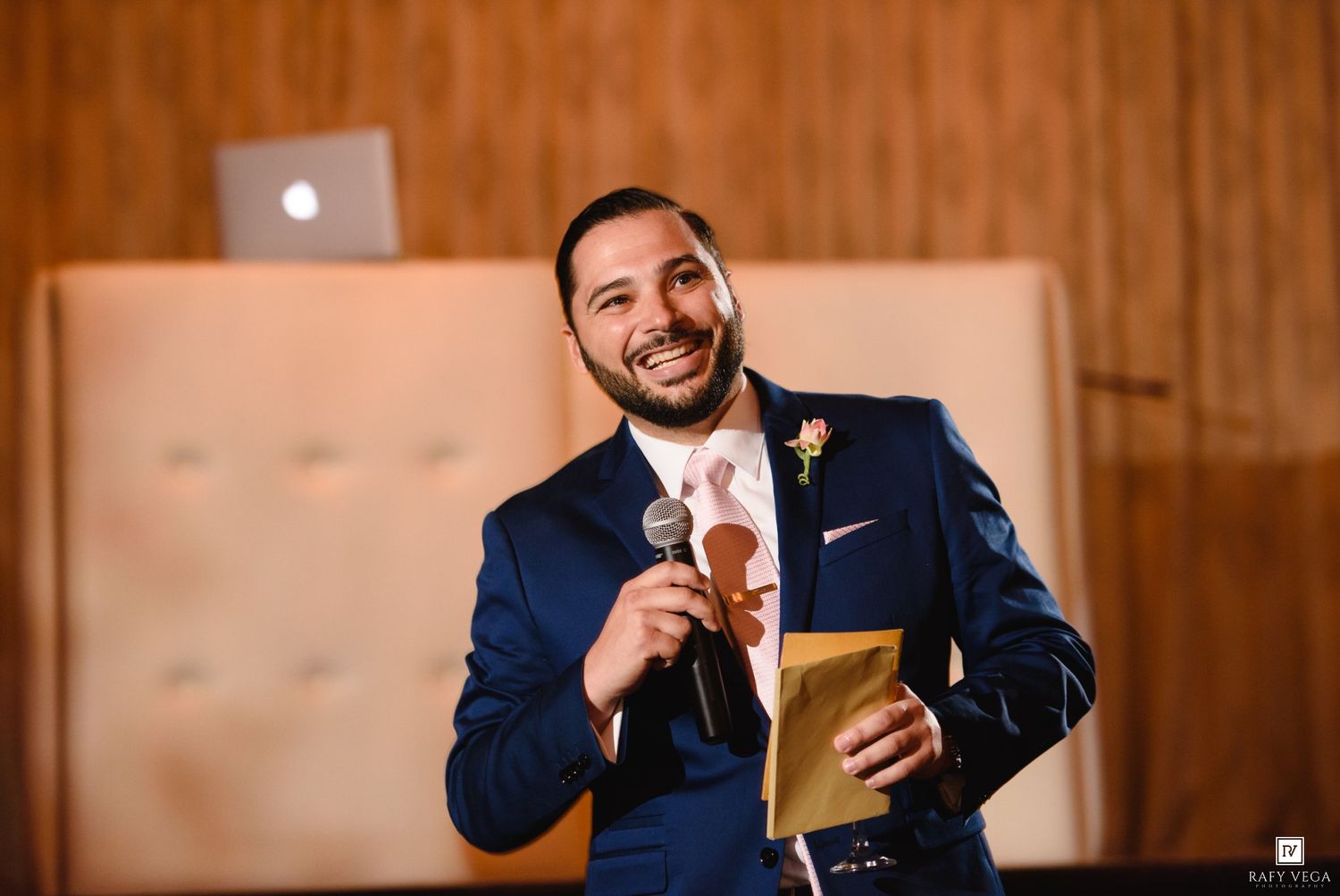 A smiling groom in a navy suit holds a microphone and envelope while giving a speech at a wedding reception.
