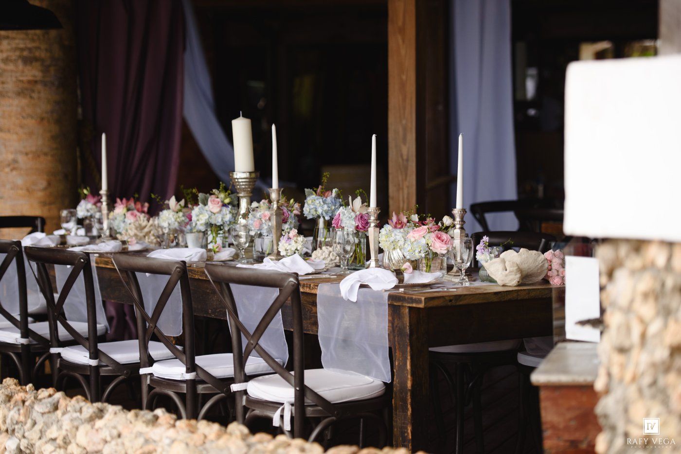 Elegant rustic wedding reception table with white candles, floral centerpieces, and dark wood cross-back chairs in a barn venue.