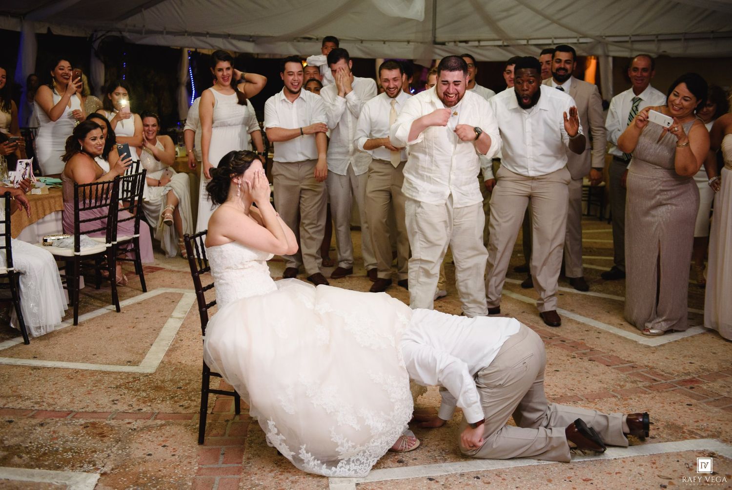 Groom retrieves garter from bride's leg at wedding reception while guests cheer and clap in background.