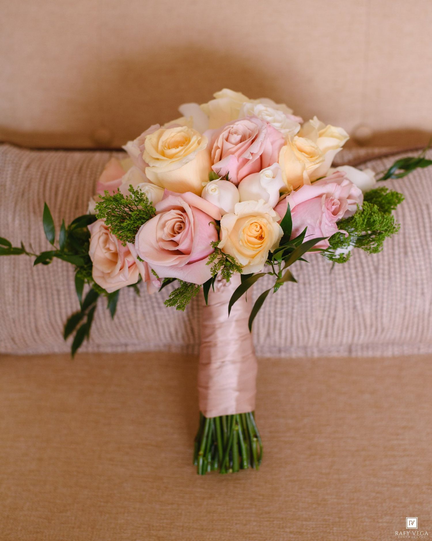 Bridal bouquet of pink and cream roses wrapped in satin ribbon resting on a beige couch.
