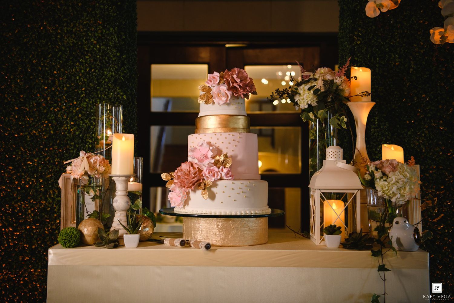 Elegant wedding cake with pink flowers and gold accents displayed on a decorated table with candles and floral arrangements.