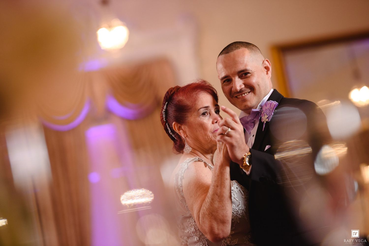 A couple shares a romantic dance at a wedding reception, with purple uplighting and elegant decor in the background.
