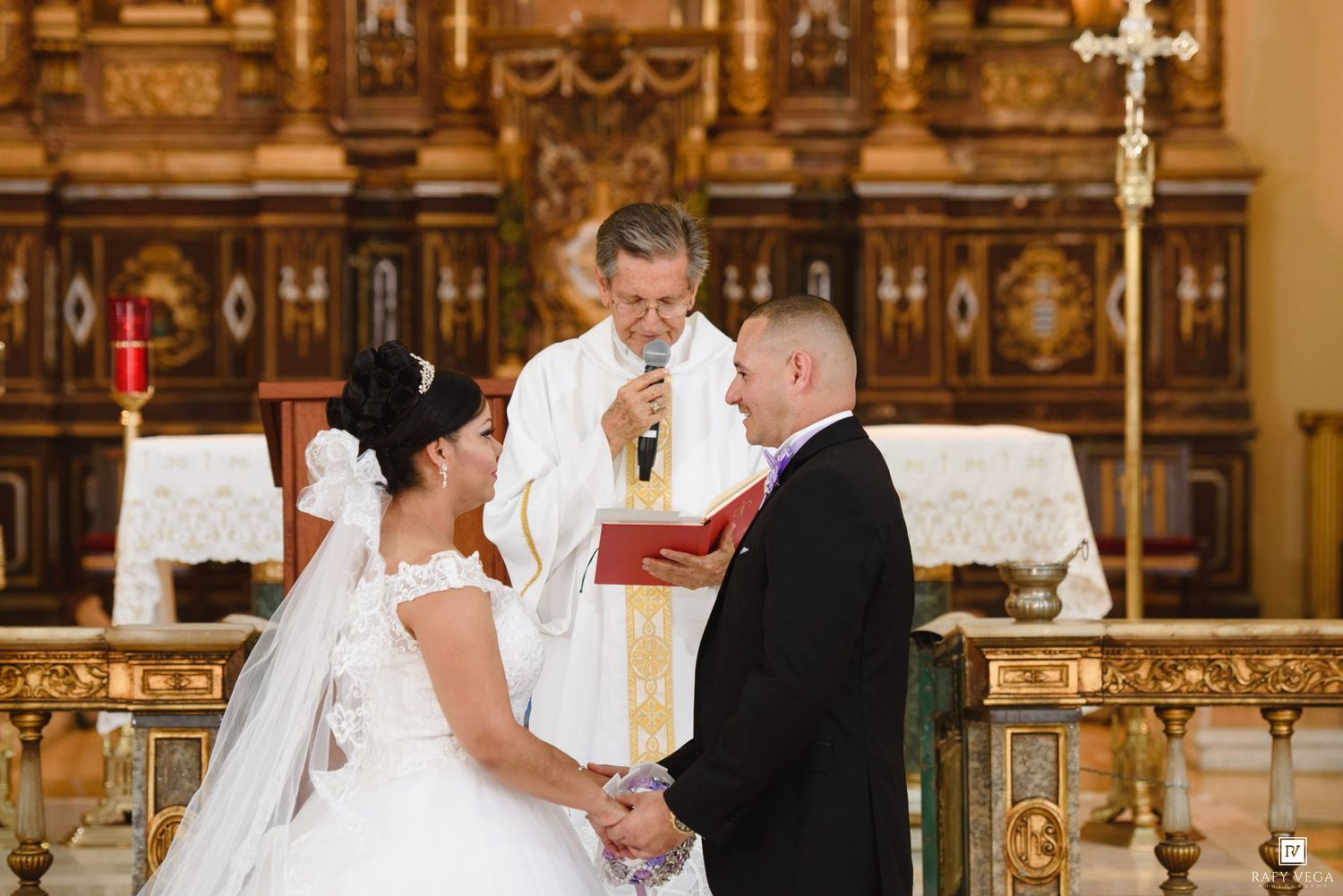 Bride and groom exchanging vows at ornate church altar with priest officiating Catholic wedding ceremony.