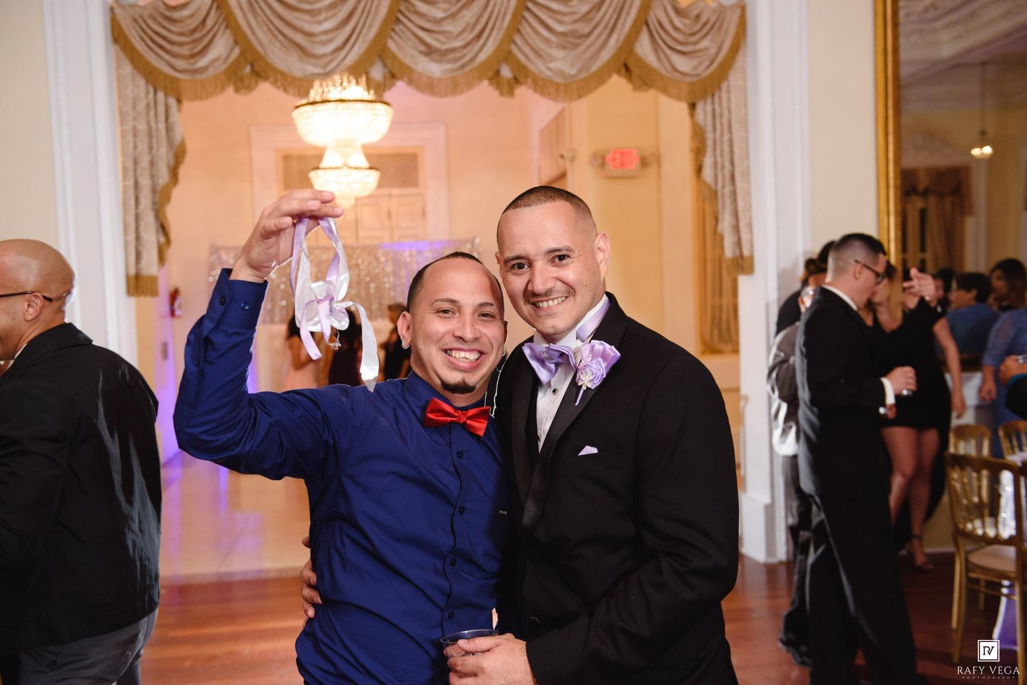 Two men smiling and holding wedding shoes at elegant ballroom reception.