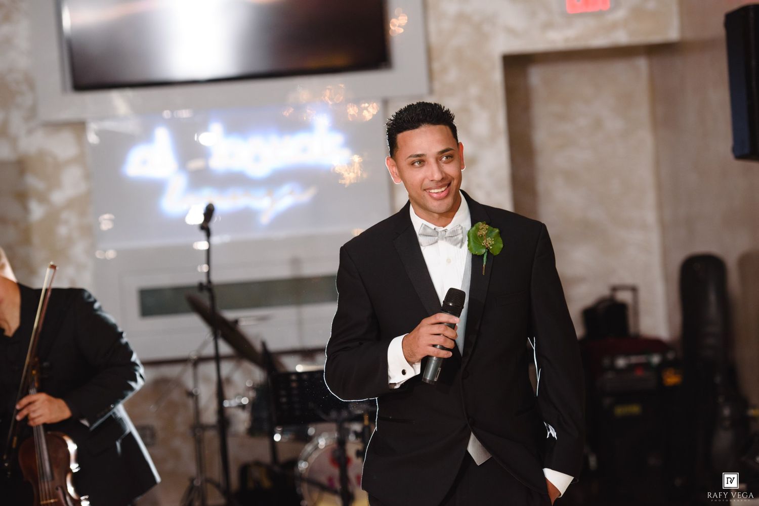 Groom in black tuxedo with white boutonniere smiling and holding microphone at elegant wedding reception.