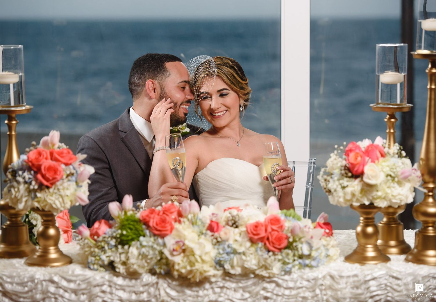 Bride and groom toasting champagne at sweetheart table adorned with coral floral centerpieces.