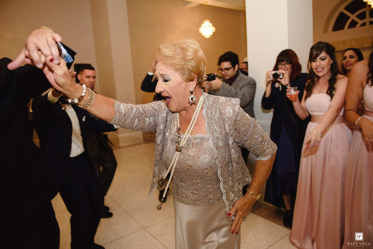 Elderly woman dancing joyfully at a wedding reception, wearing silver lace top and pearl necklace.