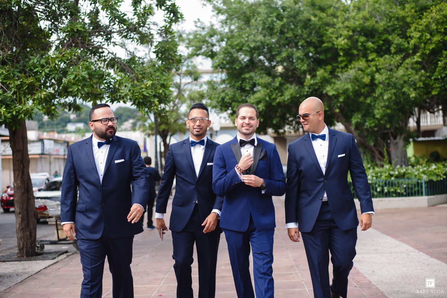 Four groomsmen in navy blue suits and bow ties walking together outdoors before a wedding ceremony.