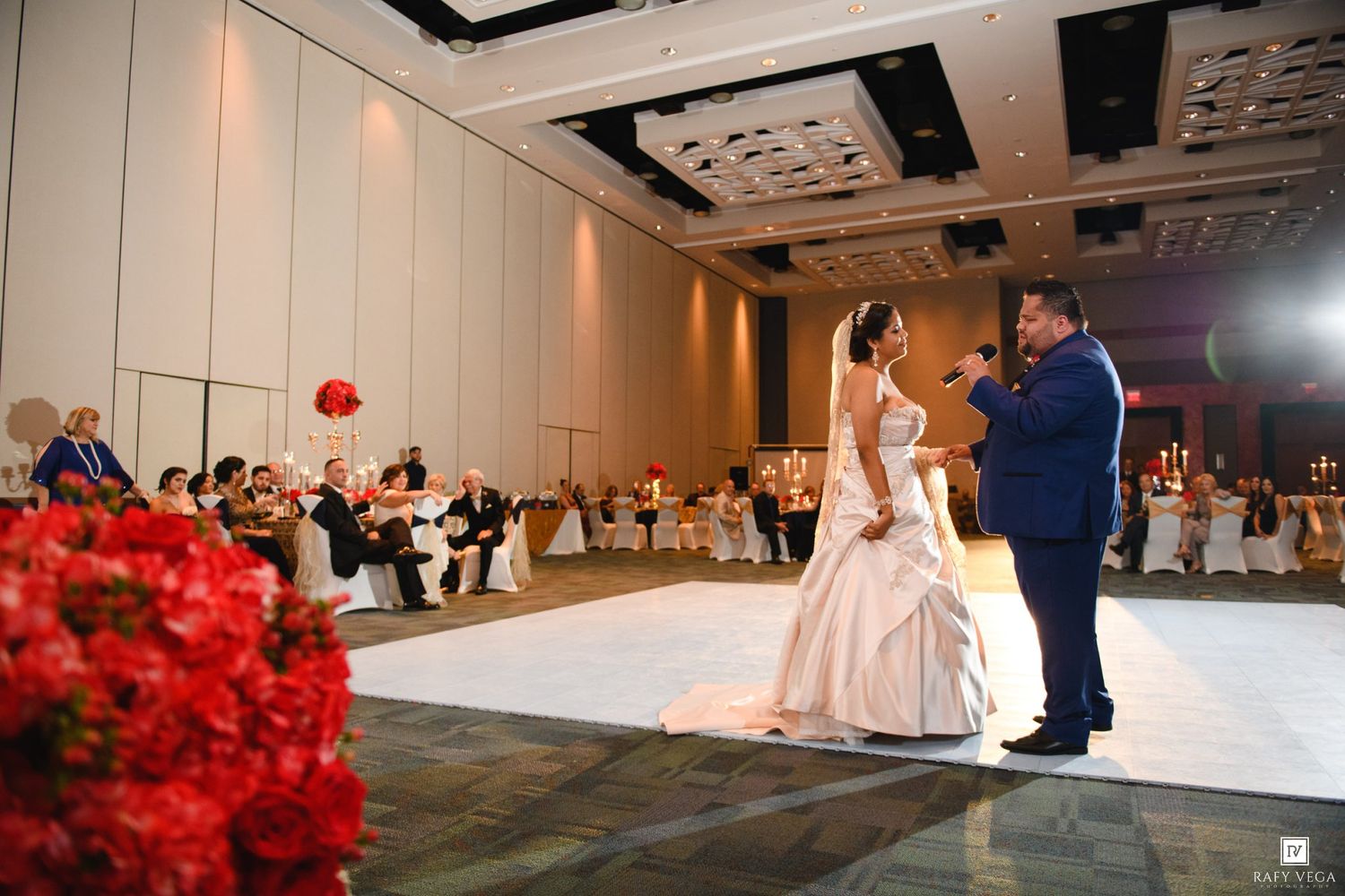 Bride and groom share first dance in elegant ballroom with red floral arrangements and seated wedding guests.