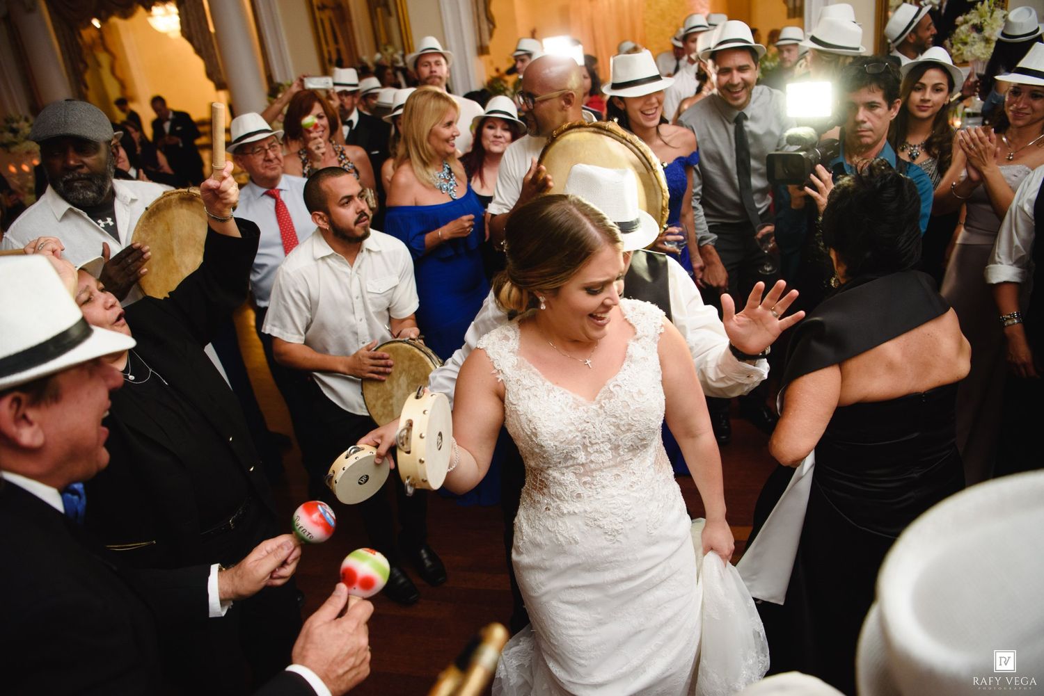 Bride dancing joyfully with guests at wedding reception, surrounded by musicians playing maracas and tambourines.
