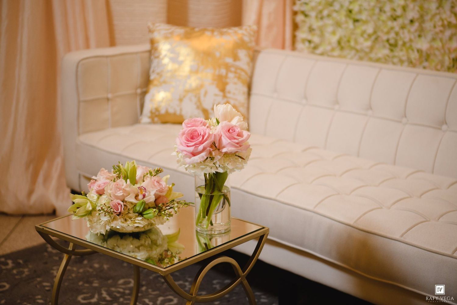 Elegant white tufted sofa with pink roses in glass vases on a gold coffee table at a wedding reception.