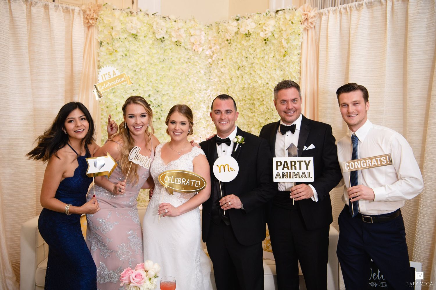 Six wedding guests smile and pose with fun photo booth props including Party Animal and Celebrate signs at a floral backdrop.