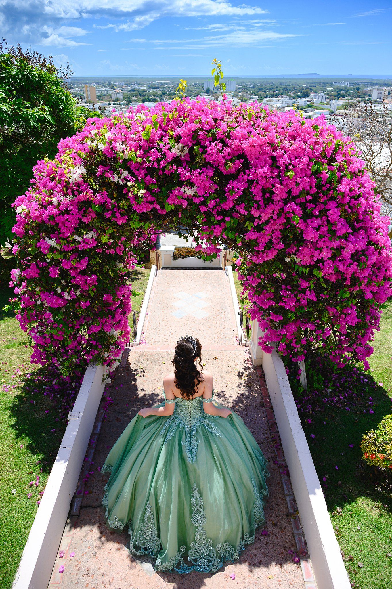 Young woman in elegant green quinceañera gown stands beneath a stunning pink bougainvillea floral arch in a garden.