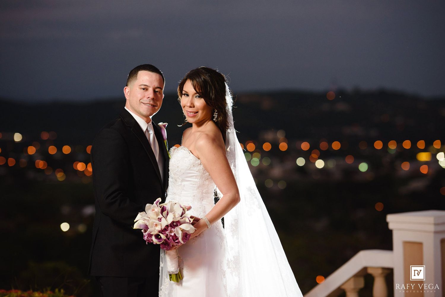 Bride and groom pose together at night with city lights bokeh backdrop, bride holding purple bouquet.