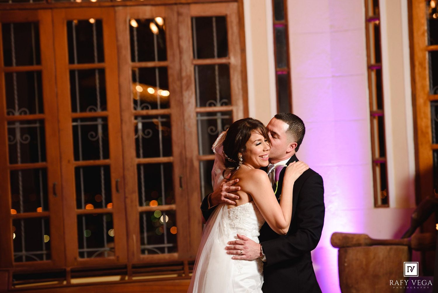 A groom kisses his bride on the cheek during their first dance at an elegant wedding reception.