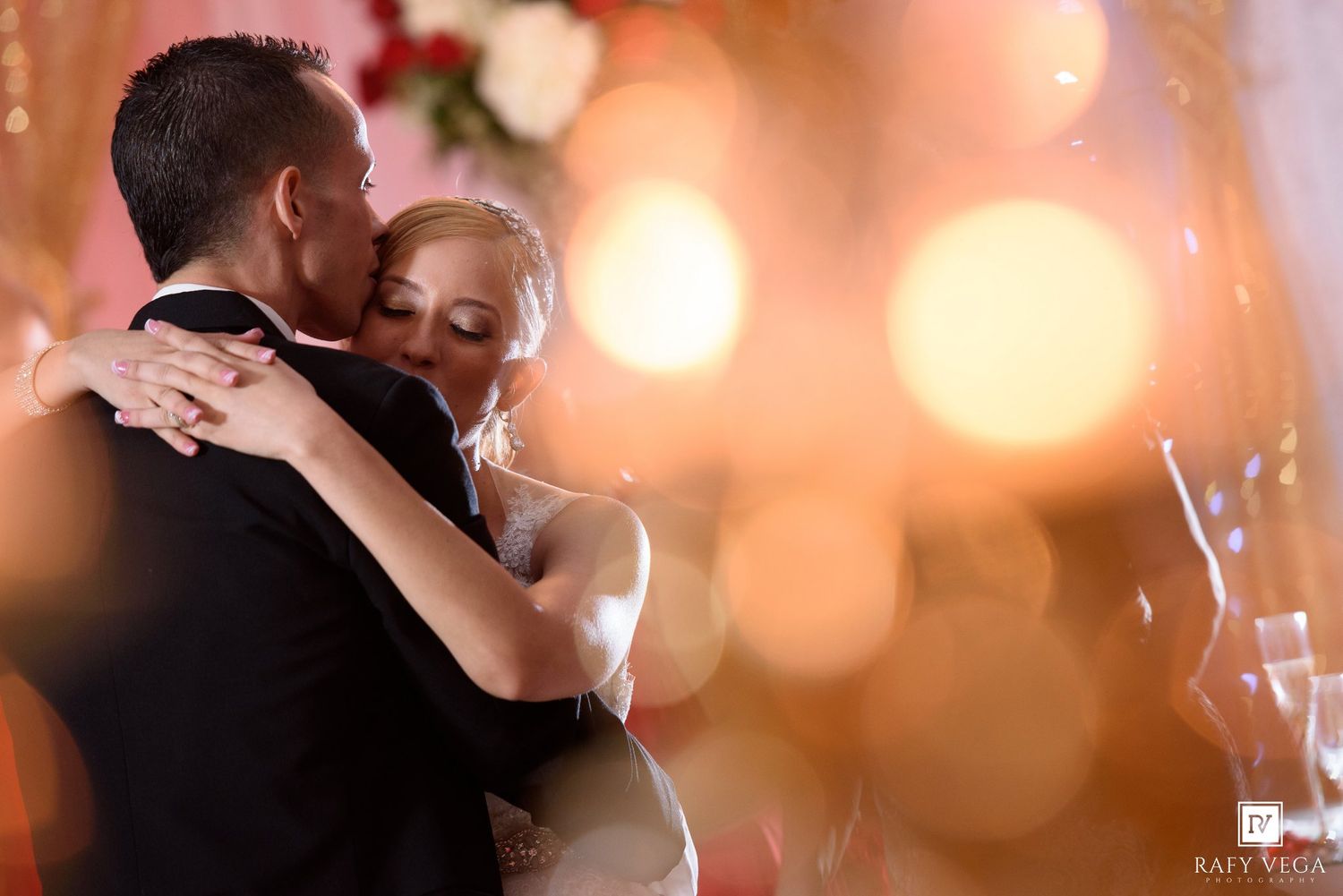 A bride and groom share an intimate first dance embrace, surrounded by warm bokeh lights at their wedding reception.