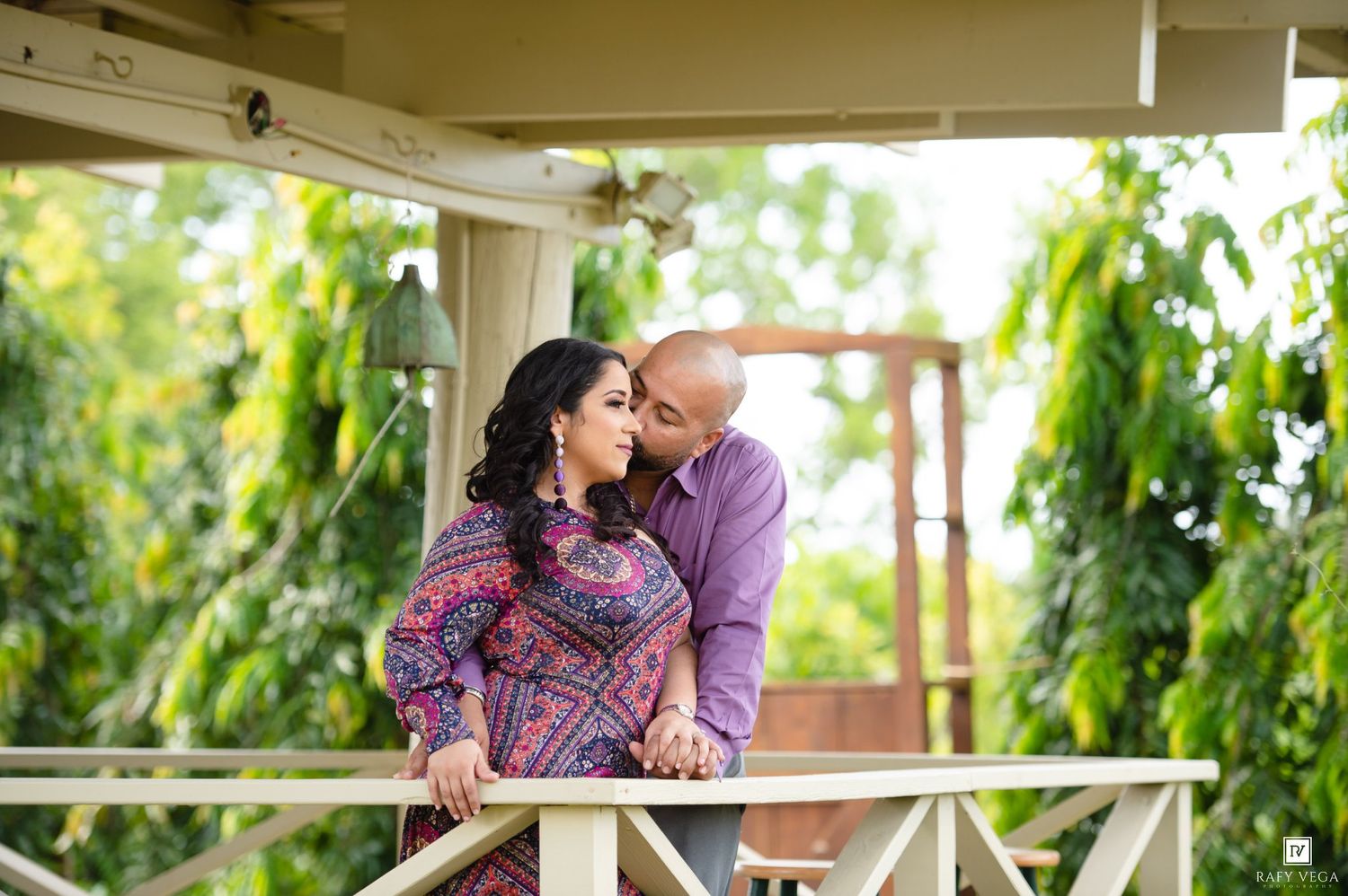 A couple shares a romantic moment on a decorated balcony surrounded by lush greenery during an outdoor engagement photoshoot.