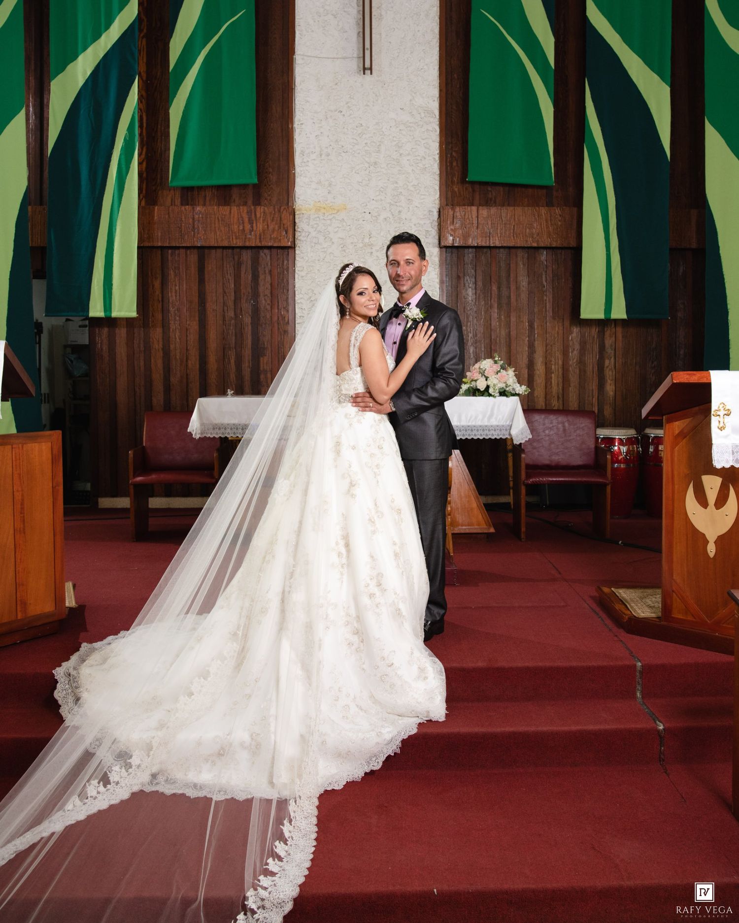 Bride and groom pose elegantly at church altar, bride wearing a stunning lace ball gown with cathedral veil.