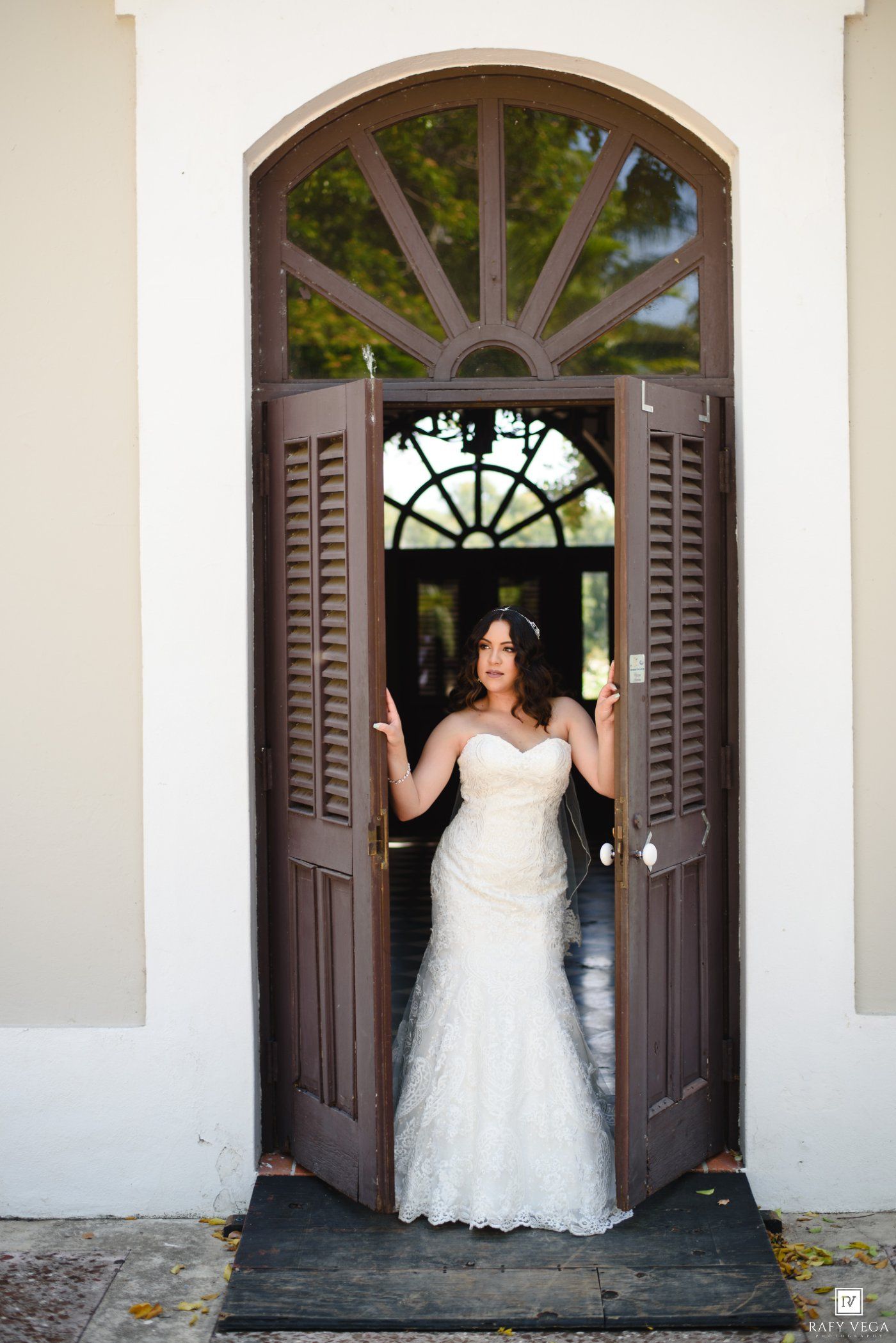 Bride in a strapless lace wedding gown standing in elegant arched double doors of a white building.
