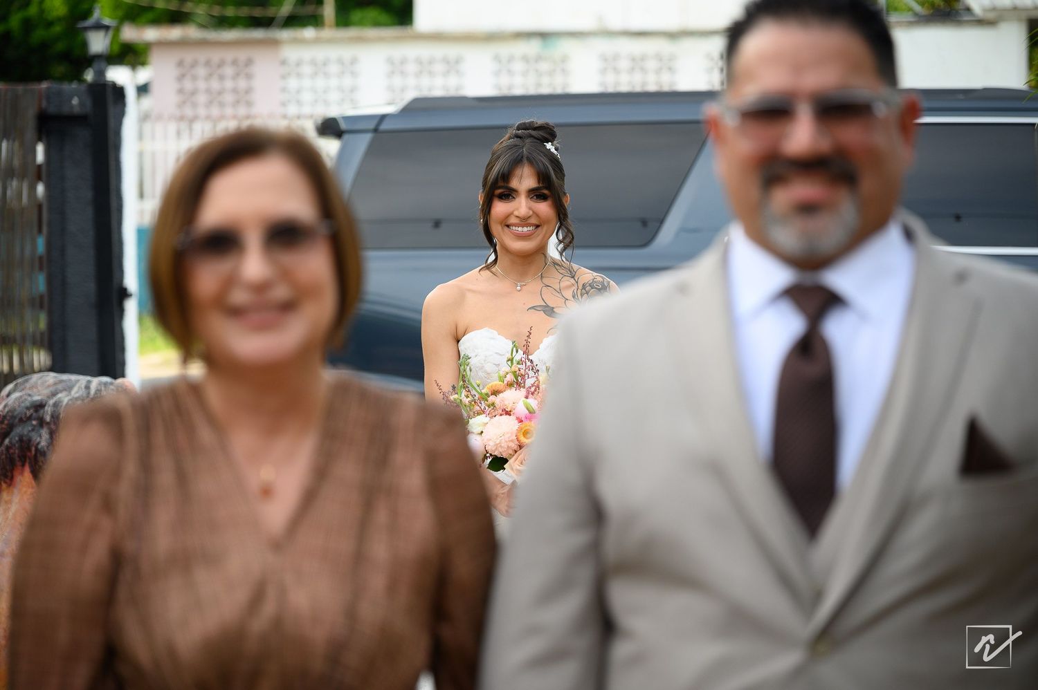 Smiling bride in a white dress holding a bouquet stands between a blurred woman in a bronze top and a man in a gray suit and tie.