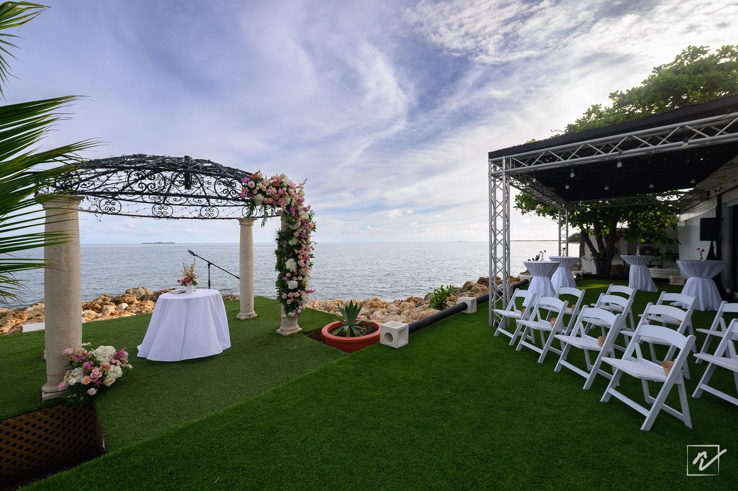 Oceanfront wedding ceremony setup with floral arch gazebo, white folding chairs, and a draped table on green lawn overlooking the sea.