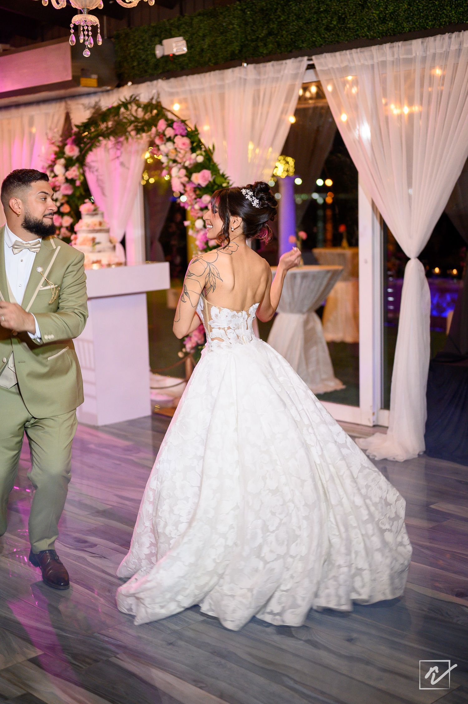 Bride in a lace ball gown and groom in an olive suit dance at an elegant wedding reception with floral arches and white drapes.