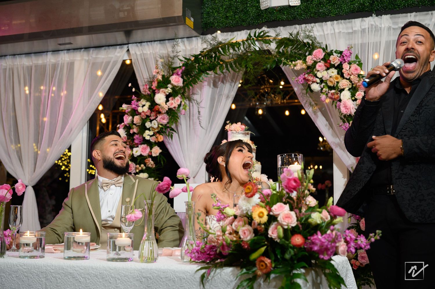 Bride and groom laugh at their wedding reception sweetheart table with pink floral arrangements as a man sings into a microphone.