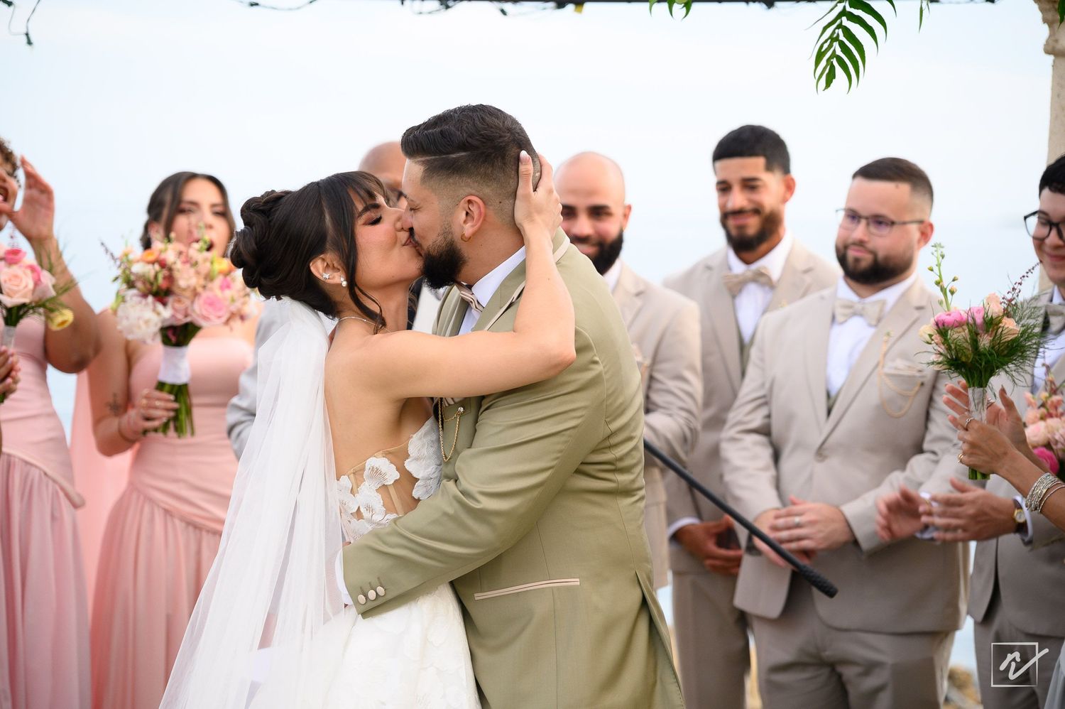 Bride in a white lace gown kisses the groom in a tan suit at an outdoor wedding ceremony as guests in pink dresses and suits cheer.
