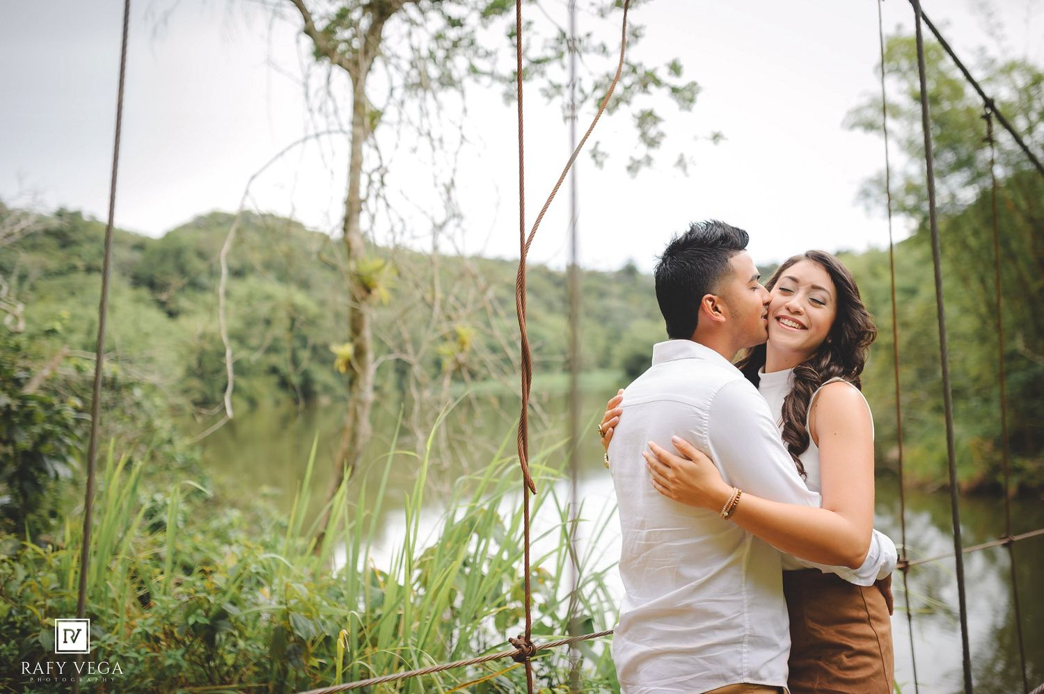 Romantic couple embracing and kissing near a lush green lake during an outdoor engagement photo session.